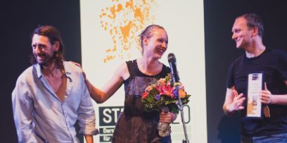 Die drei Schauspieler auf der Bühne bei der Preisverleihung. Sie in der Mitte mit Blumenstrauss in der Hand. Alle lachen.