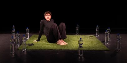 Der junge Mann trägt enganliegende schwarze Kleidung und sitzt mit angewinkelten Beinen und nachten Füssen auf einem Quadrat Kunstrasens in einem dunklen Raum. Er stützt sich auf den Händen. Um den Rasen sind einige Etikett-lose Petflaschen mit blauem Deckel aufgestellt.