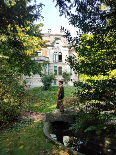 Parkanlage mit Bäumen links und rechts, sowie einem Wasser (Brunnen?), dazwischen ein Rasen, auf dem die Frau mittleren Alters steht. Im Hintergrund befindet sich das Gebäude des Literaturinstituts..