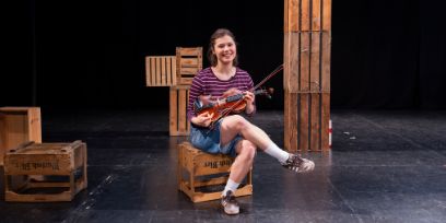 Eine junge Frau sitzt auf einer Bühne mit diversen Kisten aus Holz. Sie hat die Beine verschränkt und hält eine Violine in der Hand.