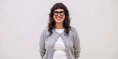 Die junge Frau sieht direkt in die Kamera und lächelt. Sie trägt ein weisses Oberteil mit eine graue Strickjacke.