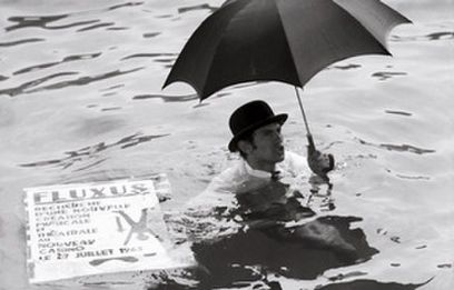 Schwarz-Weiss-Aufnahme eines Mannes im Anzug und Hut mit Regenschirm in der linken Hand. Er ist bis zur Brust im Wasser. Nehmen ihm schwimmt ein grosses Plakat auf dem "Fluxus" in in Grossbuchstaben, weitere kleinere Wörter und eine Figur im Handstand abgebildet sind.