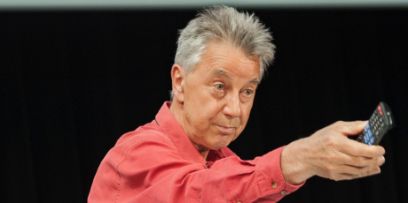Alain Findeli während einem in rotem Hemd vor schwarzem Hintergrund mit Fernbedienung in der Hand auf etwas zeigend. Oberhalb sieht man eine Projektion mit Notizen.
