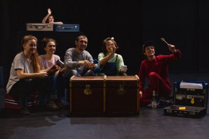 Fünf junge Menschen, davon vier Frauen und ein Mann, sitzen auf Koffern am Boden. In der Mitte steht eine grosse braune Kiste. Im Hintergrund steht eine junge Frau hinter einem Klavier und winkt.