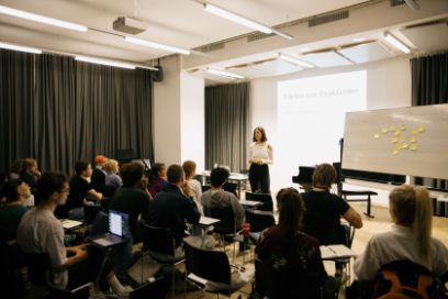 Eine Frau sthet vor einer Klasse. Im Vordergrund sind die Hinterköpfe der Studierenden zu sehen.