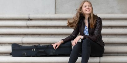 Polina Tarasenko sitzt auf einer Treppe. Sie trägt eine schwarze Lederjacke, schwarze Jeans und eine blau gemusterte Bluse. Links neben ihr liegt eine eingepackte Posaune