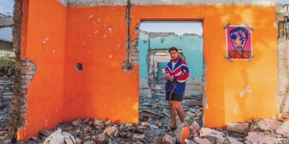 Der junge Mann steht in Shorts und Jacke auf einem Trümmerhaufen in einem Türrahmen zwischen orangefarbenen Mauern eines zertrümmerten Hauses.