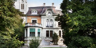 Die Aussenansicht des Standortes vom Literaturinstitut in Biel. Ein älteres, schön verziertes Gebäude steht zwischen zwei grossen Bäumen.