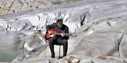 Ein Mann sitzt auf einem Stuhl und hat eine Gitarre in der Hand. Der Mann befindet sich auf einem Gletscher.