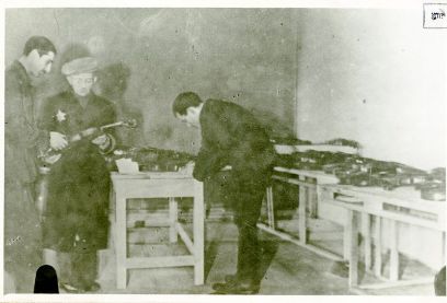 Das Schwarzweiss-Foto zeigt drei Männer, einer hat einen Judenstern am Mantel. Er und ein anderer Mann halt eine Violine in den Händen. Ein Mann rechts beugt sich über Violinen, die auf Tischen ausgelegt sind.