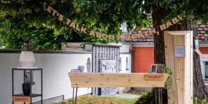 Draussen steht ein heller Holzssarg auf Beinen. Angeschrieben mit "SargBAR". Links ein Gestell, mit umfunktionierten Urnen, angeschrieben mit Jo(urn)y, rechts vor einem grossen Nadelbaum angelehnt ein weiterer Sarg.