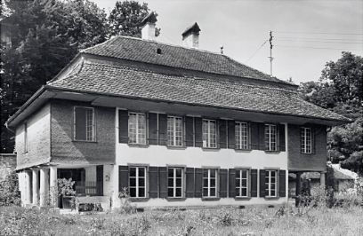 Eine Schwarzweiss-Aufnahme zeigt ein zweistöckiges Haus mit Walmdach, im Vordergrund eine Wiese, im Hintergrund Laubbäume.
