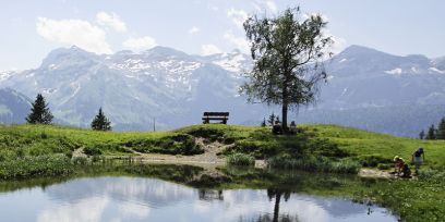 Ein kleiner Bergsee im Sommer mit einer Bank und einem Laubbaum am anderen Ufer. Am rechten Bildrand spielen Kinder, im Hintergrund sind Berge zu sehen.