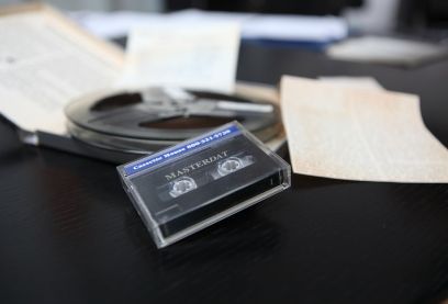 Eine Tonbandkassette liegt zusammen mit eine Tonbandspule und verschiedenen Papieren auf einem schwarzen Tisch.