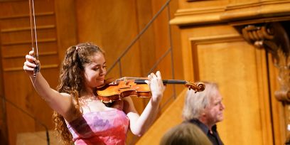 Aufnahme einer jungen Frau an der Violine vor einem holzigen Hintergrund. Sie hat lange braune Locken und ein Batikoberteil in Weiss, Violett und Pink.