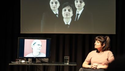Rechts im Bild sitzt eine junge Frau, auf einem Tisch links steht ein Glas Wassser und ein Bildschirm, der ein weibliches Gesicht zeigt. Darüber ist ein Bild von drei frauen auf die schwarze Wand projiziert.