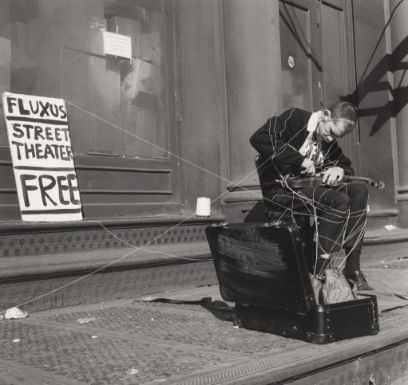 Black and white photo of a man sitting on a chair in the open, bending over a violin on his knees. The man is wrapped in strings; an open suitcase lies in the left foreground, a poster with the inscription "Fluxus Street Theater Free" hangs in the left background.