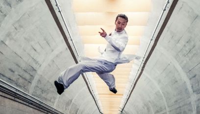 Ein asiatischer Kung-Fu-Kämper in weiten Hosen und einem weissen Hemd springt in einem hell ausgeleuchteten, bentonierten Tunnel mit gespreizten Beinen hoch in die Luft.