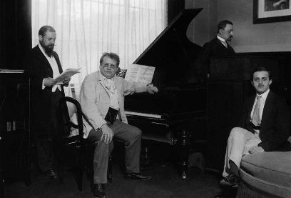 Schwarzweiss-Fotografie von vier Männer. Der erste von links steht und schaut in ein Notenblatt, der zweite von links sitzt am Flügel und schaut in die Kamera, der dritte steht mit dem Rücken zum Pianisten, der vierte sitzt auf einem Sofa und schaut in die Kamera.