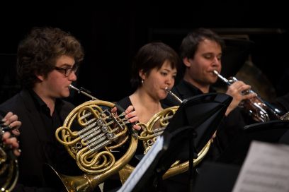 Ein Hornist und eine Hornistin, dahinter ein Trompeter, sitzen im Orchester und spielen.