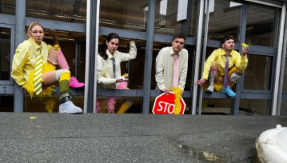Vier Schauspieler sitzen im Fenster mit gelben Kleidern, einer hält ein Stoppschild, vorne Betonboden