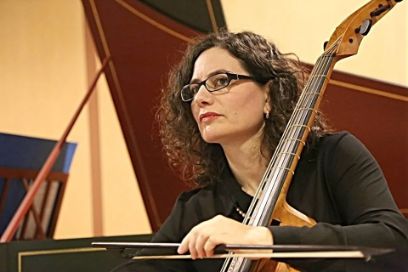 Eine Frau mit langen Haaren und Brille sitzt, ein Cello im linken Arm, vor einem Cembalo.