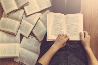 Eine auf dem Fussboden liegende Person ist von oben Fotografiert mit einem Buch in den Händen, links liegen zahlreiche aufgeschlagene Bücher.