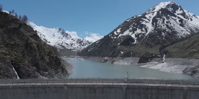 Foto eines teilweise gefüllten Stausees, im Vordergrund die Krone der Staumauer, im Hintergrund schneebedeckte Berge.