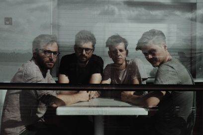 Foto in Grautönen: vier Männer an kleinem Tisch. Durch ein Glas fotografiert, Wolken spiegeln sich darin. Die Männer tragen Kurzarm-Shirts und ihre Haare kurz.