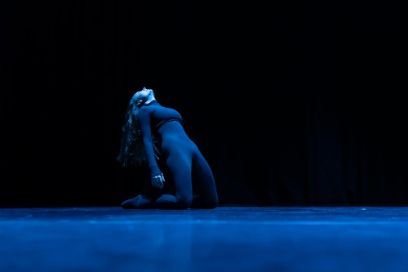 Une femme éclairée en bleu se penche en arrière, à genoux. L'arrière-plan est noir.