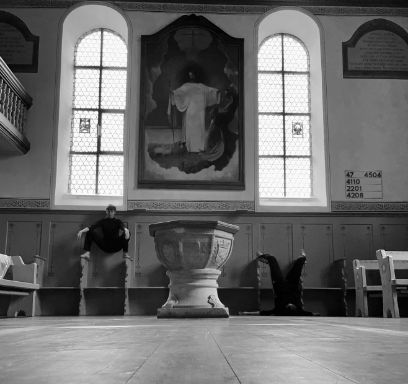 Schwarzweissaufnahme des Chors einer Kirche mit Taufstein im Vordergrund und Chorgestühl im Hintergrund. Im Chorgestühl sind zwei Personen zu sehen.