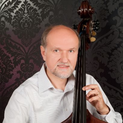 Ein Mann in weissem Hemd vor dunklem Hintergrund hält einen Kontrabass und schaut gerade in die Kamera.