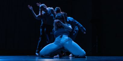 Foto mit Tänzer*innen in dunklem Raum, Vorderste auf Knien, Personen blau beleuchtet