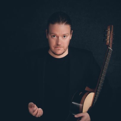 Ein Mann in schwarzem Pullover vor schwarzem Hintergrund hält eine Gitarre im linken Arm und schaut gerade in die Kamera.