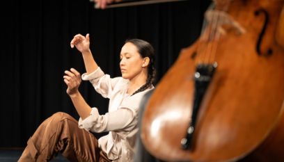 Cello im Vordergrund (verschwommen), dahinter eine Performerin, die mit den Händen sanfte Gesten ausführt
