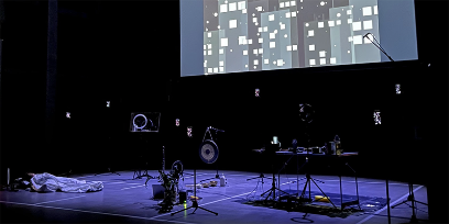 Blick auf eine Bühne, am Boden liegt eine Person, rundherum diverse Gegenstände auf Stativen. im Hintergrund eine grosse Leinwand