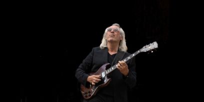 Ein Mann mit Brille und schulterlangen grauen Haaren spielt vor schwarzem Hintergrund elektrische Gitarre.