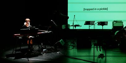 Eine Person an einem Synthisizer auf dunkler Bühne, im Hintergrund ein Screen in grün