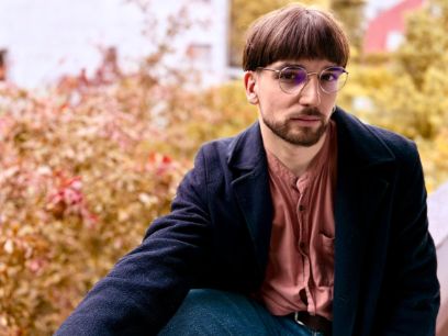Ein junger Mann mit Brille und Bart sitzt im Freien auf einer niedrigen Mauer und blickt in Dreiviertelansicht in die Kamera.