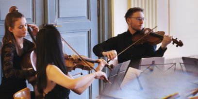 Eine Cellistin, eine Geigerin und ein Geiger sitzen hinter Notenständern und spielen ihre Instrumente.