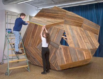 Ein Mann auf einer Leiter und eine stehende Frau halten ein Messband an eine Holzskulptur.