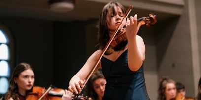 Im Vordergrund eine spielende Violinistin, im Hintergrund Streicherinnen, die sie begleiten.