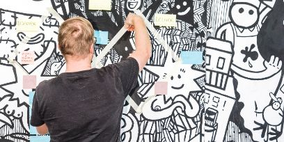 Junger Mann in schwarzem T-Shirt vor einer Wand voller Skizzen mit Post-it und Klebeband am Brainstorming machen.