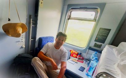 René R. Schmidpeter sitzt in einem Einzelabteil eines Nachtzugs. Rechts sieht man das Bett, links sitzt er auf einem Sitz und lacht in die Kamera.