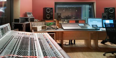 Studio d'enregistrement avec des vitre sur la gauche et dans le font de l'image. Le mure est dans un rouge/orange, les meubles et le sol est en bois claire.
